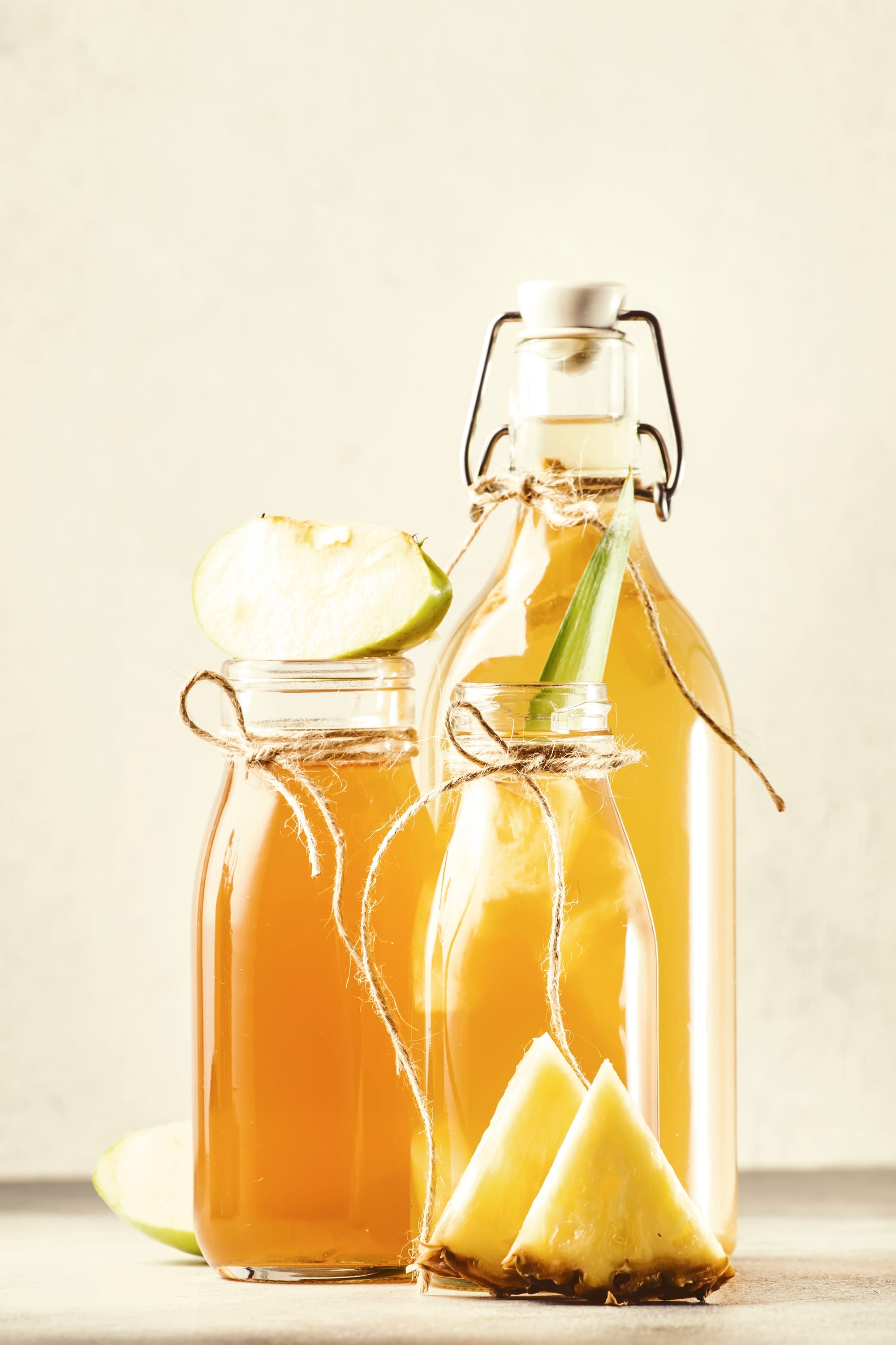 Side view of three bottles of tepache on an off-white background. There are two pieces of pineapple in front of the bottle. The bottles have string around the necks. One of the bottles also has a lid.