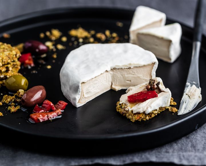 Vegan camembert cut open on a black plate with crackers, olives and peppers in olive oil.