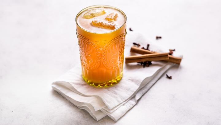 Glass of tepache with ice cubes on a white napkin with cloves and cinnamon sticks on the white tabl