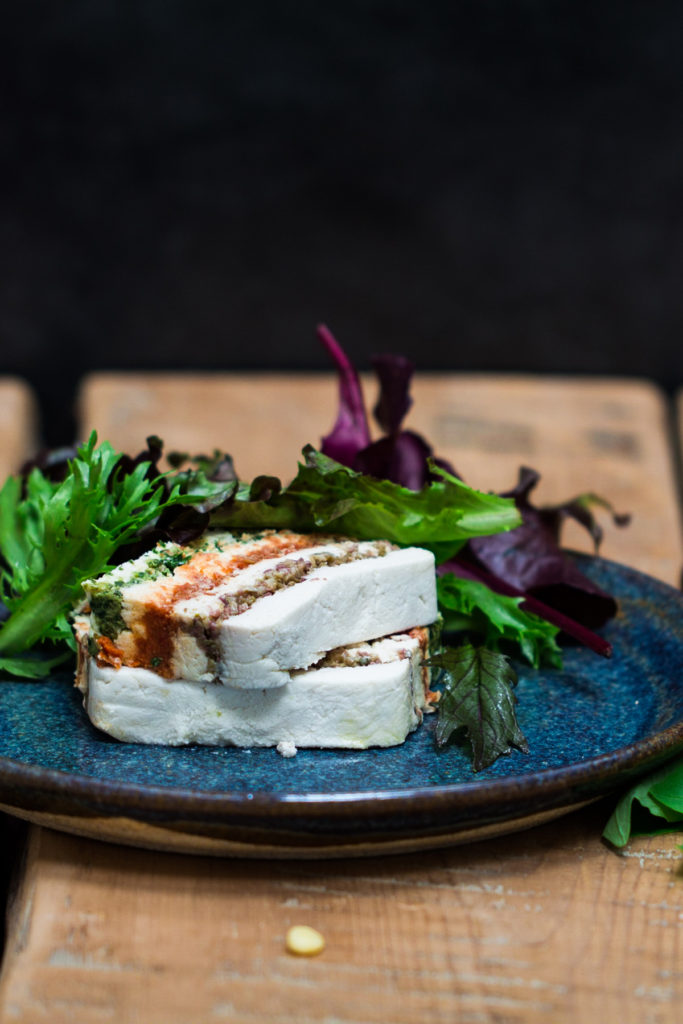 The Almond Cheese Terrine is stacked into two layers with salad greens on the side. The dish is served in a blue plate on a wooden table.