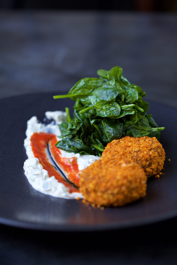 There are two croquettes served in a gray plate with the tartar and sweet chilli spread accross. There is also the wilted spinach on the side of the croquettes.