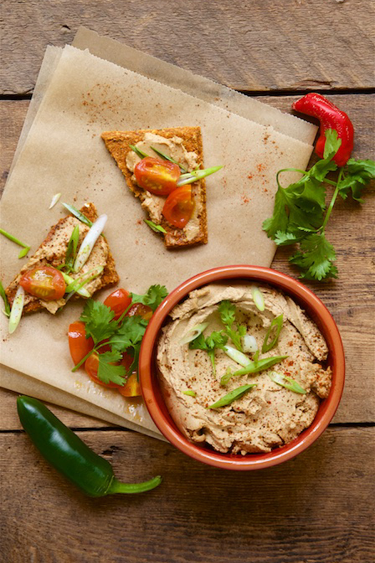 The dip is in a colored bowl with some vegan pizza on top of a table napkin. Some red and green chillies are also present on the table.