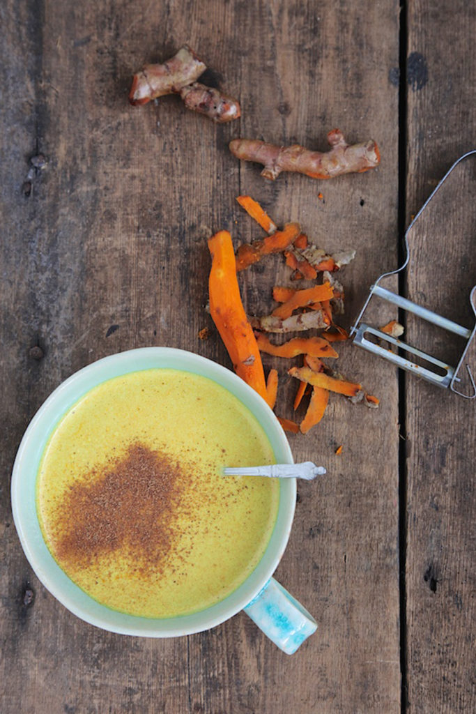 The tea is in a white cup with the some the turmeric roots on top of a wooden table with the peeler visible on the side.