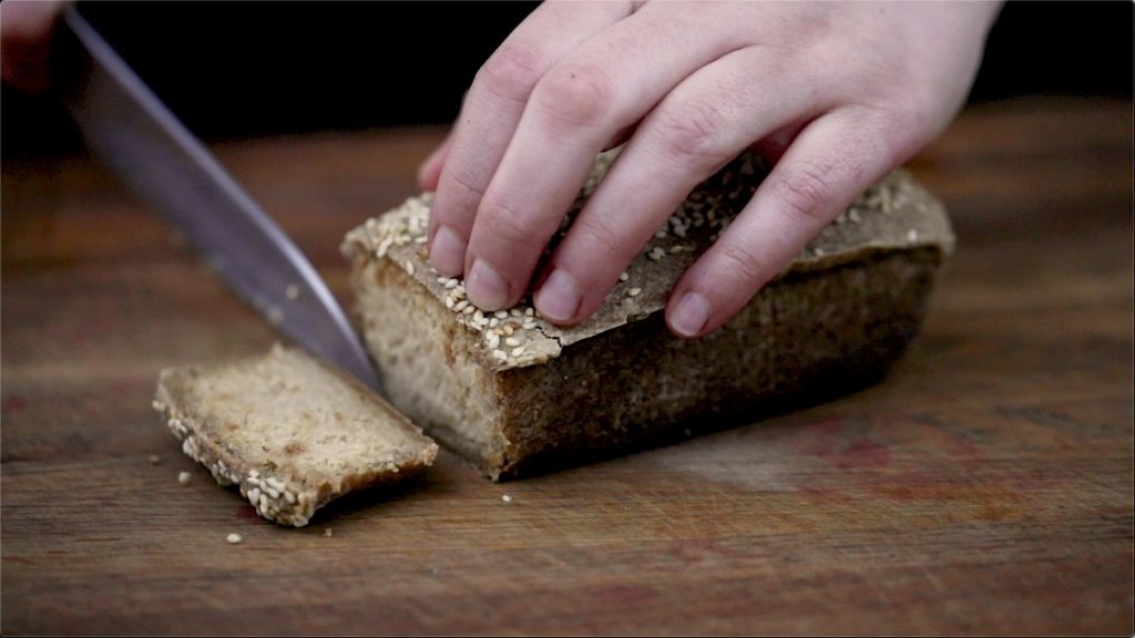 Fermented raw vegan bread loaf with one slice being cut off on a wooden chopping board