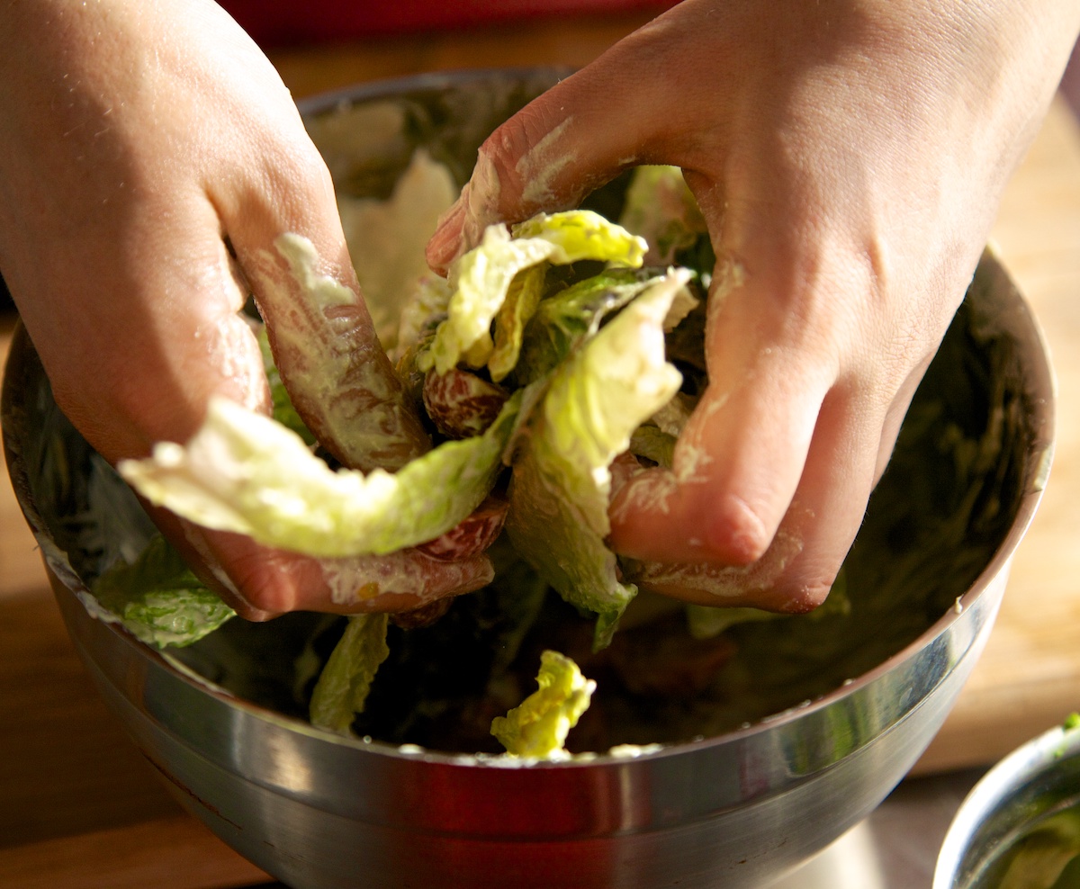 hands mixing a raw vegan caesar salad dressing with romaine lettuce leaves in a silver bowl