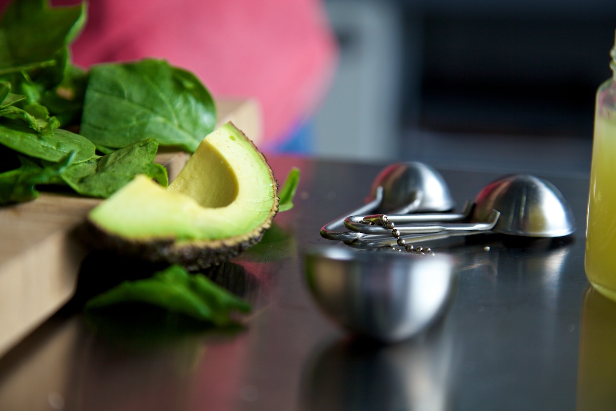 Quarter of an avocado, raw spinach, jar of lemon juice and measuring spoons on a stainless steel surface.